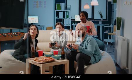 Diverse group of workmates using joysticks to play video games on television. Colleagues cheering while playing game with controllers on tv console to have fun and entertainment. Stock Photo