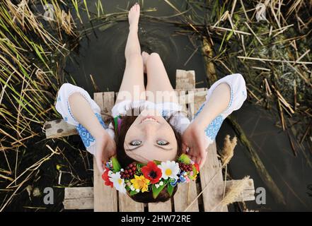 girl in a flower wreath on his head sitting on the bridge and wets feet in the river Stock Photo