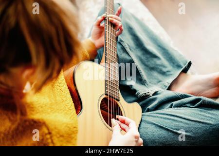 Young girl with long nails plays acoustic guitar at home. Teenager sits on couch in room and learns to play musical instrument. Background. Close-up. Stock Photo