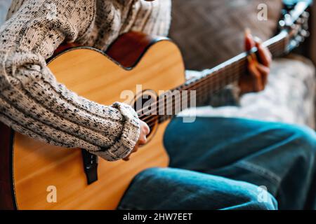 Young girl with long nails plays acoustic guitar at home. Teenager sits on couch in room and learns to play musical instrument. Background. Close-up. Stock Photo