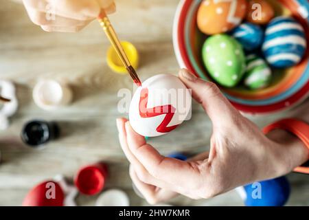 Woman's hand with a brush is drawing a colored pattern on an Easter egg. Creative preparation for bright Easter holiday Stock Photo