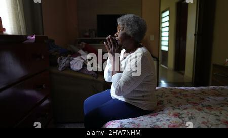 A spiritual older black woman praying to God sitting in bedroom Stock Photo