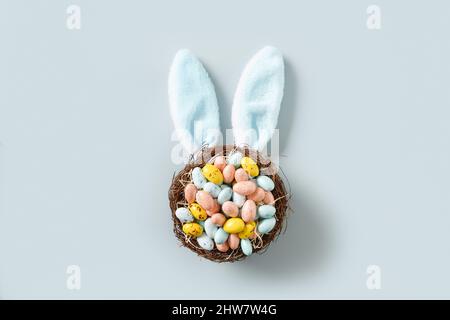 Easter nest with pastel chocolate eggs decorated funny bunny ears on blue background. View from above. Flat lay minimal style. Happy Easter greeting c Stock Photo