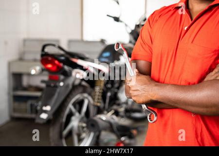 Close up shot of mechanic with tool in hand standing with arms crossed in front of bike at garage - concept of repair and maintenance service showing Stock Photo