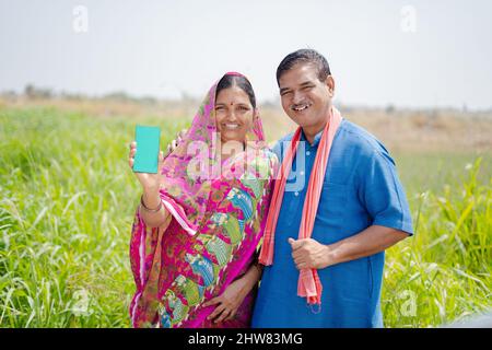 focus on man, happy village couples showing green screen mobile phone by looking at camera at meadow - concept of advertisement, app or application Stock Photo