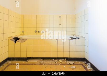 The bathroom is broken. Pieces of ceramic tiles and garbage on the floor. The water plug hangs on the wall. Stock Photo