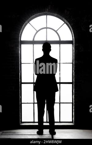 A silhouette of a man in a suit with his arms crossed facing an arched window with the light streaming in illustrating a decision, power and the Stock Photo
