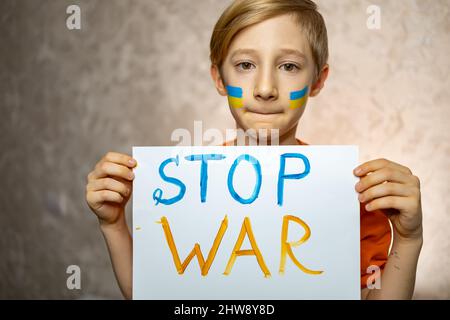 a child against war, a boy with a painted flag of Ukraine on his cheeks holding a poster with the inscription stop the war Stock Photo