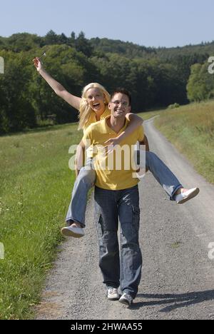 man walking along a country lane and carrying his wife piggyback, Germany Stock Photo