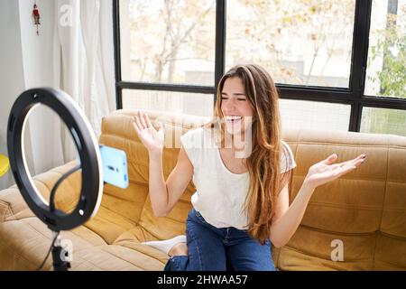 Modern influencer. Female vlogger making social media video while sitting indoors sofa home shooting with mobile smart phone camera. Stock Photo