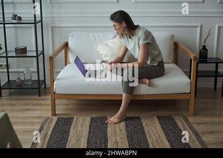 Young happy woman using laptop, typing and using touchpad, chatting online, drinking coffee, sitting on sofa at home Stock Photo