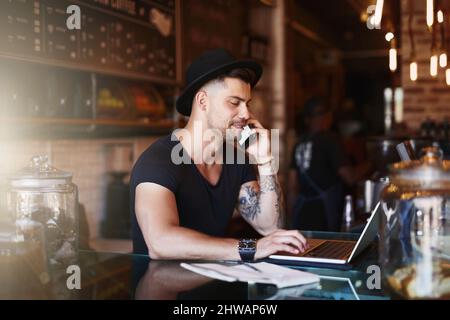 Busy behind the bar. Shot of a young man using a phone and laptop while working in a coffee shop. Stock Photo