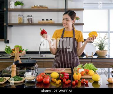 Young housewife stand smiling,  hold red and yellow bell pepper with both hands. Looking at the red one on the right. The kitchen counter full of vari Stock Photo