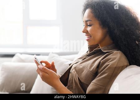 Side view at young multiracial woman using trendy smartphone sitting on the sofa at home, ethnic female chatting in social medias, texting, scrolling news feed, sharing photos, using mobile app Stock Photo