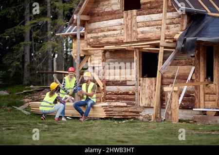 Young construction workers chatting in front of wooden house. Wooden Skeleton Framing Building. Construction industry, real estate, teamwork concept. Stock Photo