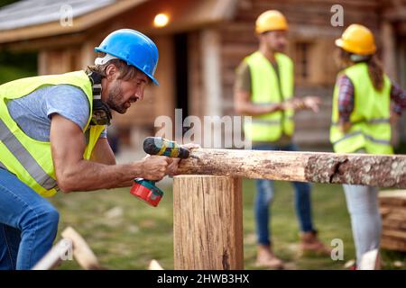 Young constructer worker, working with drilling machine on wooden construction. Other workers having a conversation in the background. Wooden Skeleton Stock Photo