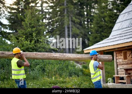 Two young male construction workers carrying big heavy wood on a construction site, building a forest wooden house. Wooden Skeleton Framing Building. Stock Photo