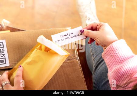 High angle of crop anonymous female sender packing order in envelope and putting note thank you for purchase before shipping in box Stock Photo