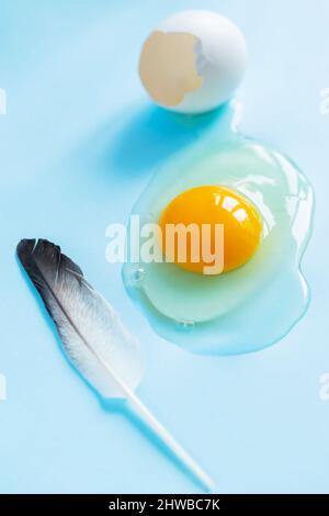 Broken chicken egg and feather on blue background. Broken eggshell with liquids inside. Easter concept Stock Photo