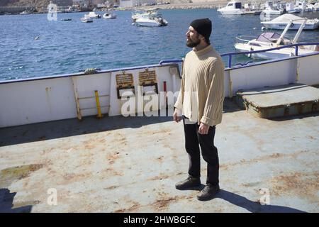 Tall and robust young sailor man with a beard in a woolen cap and high-necked pullover is standing on a ship in a sea port Stock Photo