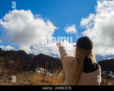 The girl points her finger at the white peak of Elbrus, visible through the clouds against the background of bright blue sky, standing in an autumn mo Stock Photo