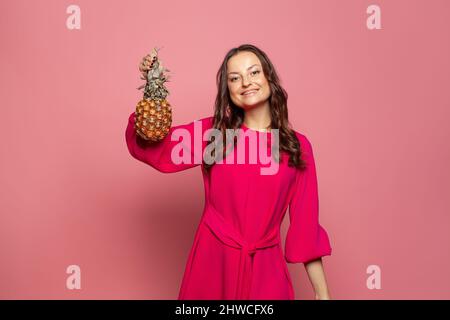 Smiling woman in red summer dress holding pineapple in her hand and smiling on pink background Stock Photo