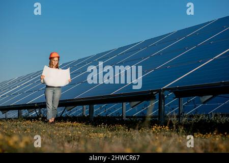 Handsome Architect Woman Examining a Draft Map or Blueprint Project Plan, Worker Activity Looking Out in Photovoltaic Cell Farm or Solar Panels Field. Stock Photo