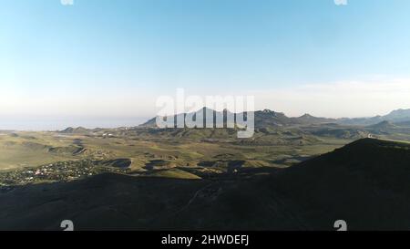 Aerial view of the small settlement in green hills with high mountain chain and blue sea on the background against blue sky. Beautiful colorful landsc Stock Photo