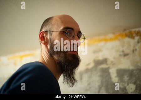 Man is thinking about repairs. Guy with glasses against background of wall. My husband remembers how to glue wallpaper. Face with long beard. Stock Photo