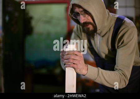 Guy works on wood. Carpenter holds board. Man makes furniture. Man in carpentry workshop. Man in garage. Stock Photo