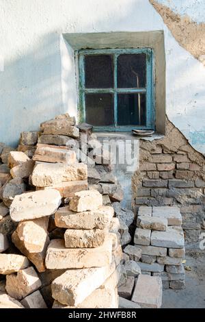 Small framed dusty window in a wall with disintegrated plastering showing layers of сement and brick and stacks of bricks Stock Photo