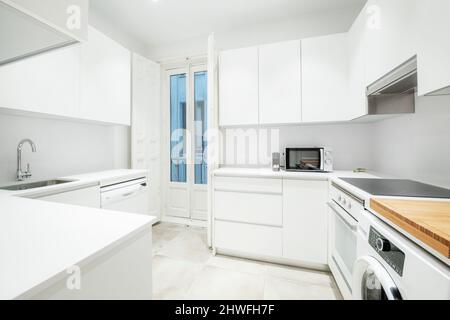 Nice kitchen of a short-term rental apartment with white furniture and appliances, with a balcony with vintage wooden doors and a ceramic hob Stock Photo