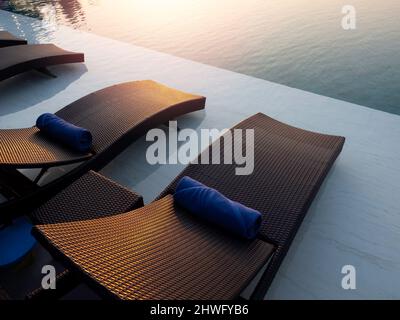 Two empty modern rattan sunbeds, chairs decorated with blue rolled towels prepared for the guest on white marble floor at swimming pool in a hotel res Stock Photo