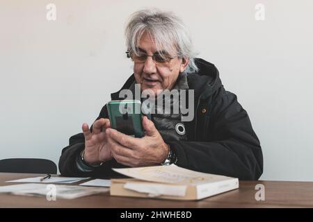 Happy stylish mature old man remote sitting at the desk, using a smartphone. Books, paper and a pen on the desk. Smiling 60s middle aged man. Stock Photo