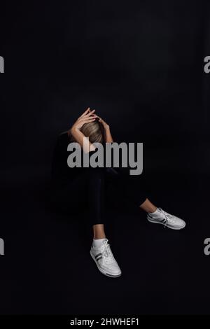 Depressed Caucasian female in black dress sitting on floor hiding face with arms in despair with black background. Mental and physical abuse. Rough Stock Photo