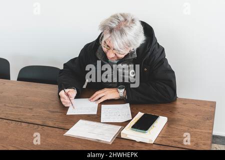 Happy stylish mature old man sitting at the desk, writing on paper. 60s middle aged man. Stock Photo