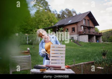 Senior woman with paintbrush impregnating wooden crate outdoors in garden. Stock Photo