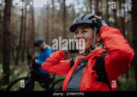 Senior couple bikers putting on cycling helmet outdoors in forest in autumn day. Stock Photo