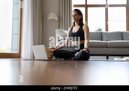 Woman do meditation practice seated cross-legged on mat near laptop Stock Photo