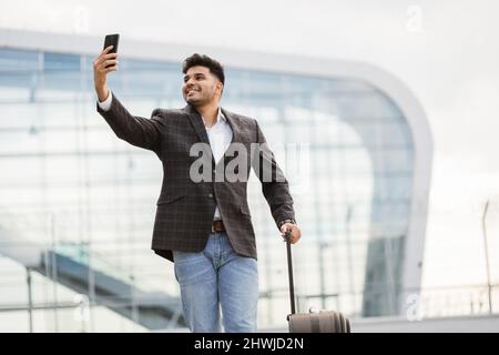 People, lifestyle, travel, tourism and modern technology. Handsome happy Indian businessman making video call on smartphone to his friend and showing hello gesture against modern airport background Stock Photo