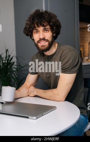 Vertical portrait of cheerful curly young businessman holding in hand cup with morning coffee sitting at table with closed laptop computer Stock Photo