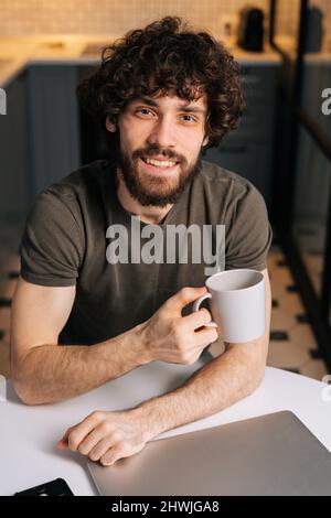 High-angle view of smiling bearded young business man holding in hand cup with morning coffee sitting at table with closed laptop computer Stock Photo