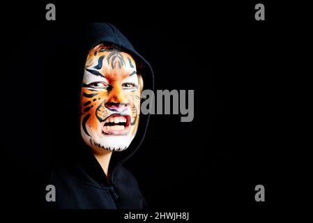 Portrait of a boy with strange and scary gesture, made up with tiger face, isolated on black background. Stock Photo