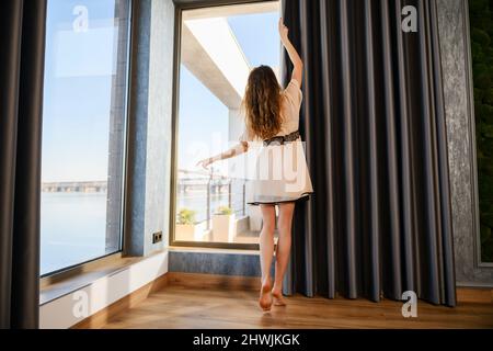 A young attractive girl opens the curtains in the morning. Back view. The woman is dressed in a peignoir. Stock Photo