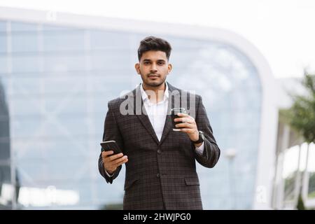 Handsome Indian businessman in formal wear, holding a cup of coffee looking away. Young thoughtful Arab man standing outside modern building and drinking take away coffee Stock Photo