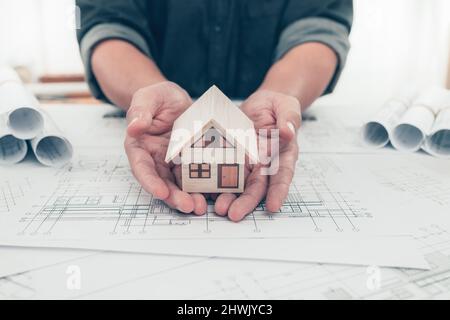 Architect engineer hands hold and protect mini house model on bueprint. Protect House, cost planning design and construction concept. Stock Photo