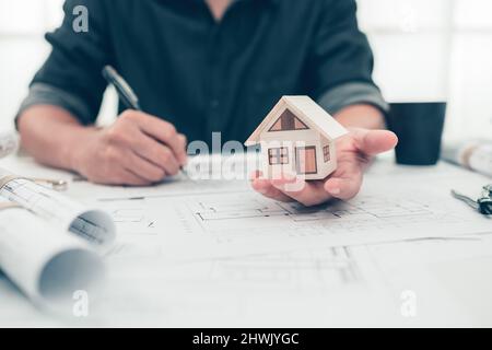 Architect engineer hands hold and show mini house model and use pen drawing on bueprint. House planning design and construction concept. Stock Photo