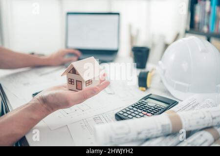 Architect engineer hands hold and show mini house model and use laptop on desk. House planning design and construction concept. Stock Photo