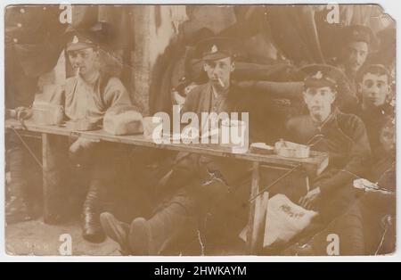 Informal photograph of British First World War soldiers eating rations. The young men are sitting at a low bench or table which has bread, mess tins & tin mugs on. Two are smoking cigarettes & one has a newspaper on his lap, one soldier looks as though he's wearing a cardigan, in addition to his standard army uniform. The accommodation looks temporary, perhaps a barn or other building on the Western Front Stock Photo