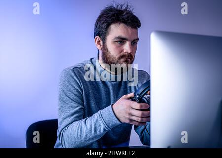A man sits in front of a computer, holding headphones in his hands. Stock Photo